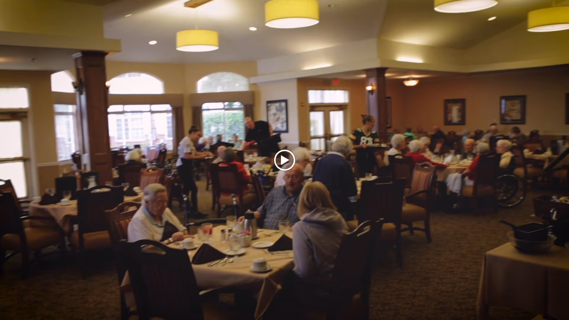 Large dining room in a senior living facility with many residents seated at tables while staff serve meals.