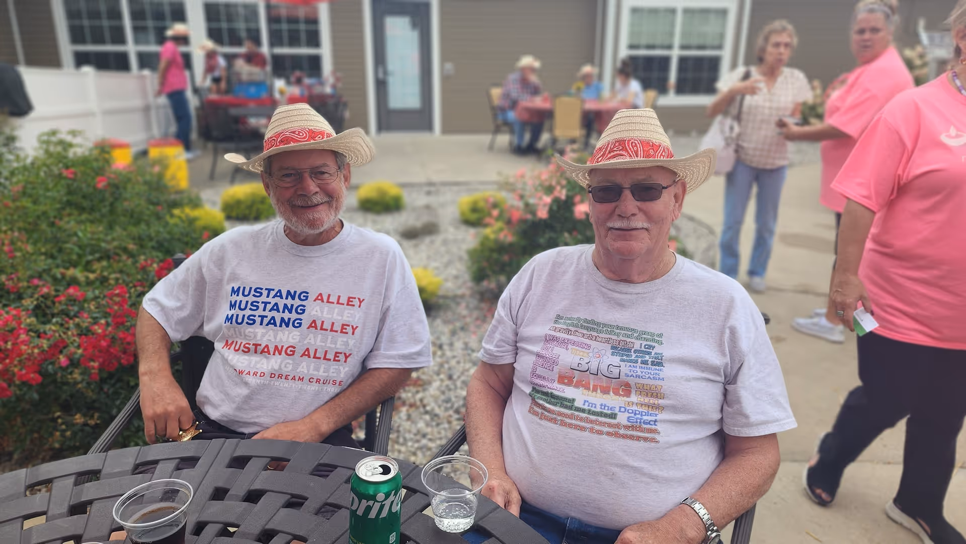 Two elderly men wearing straw hats and casual t-shirts sitting at a round outdoor table with drinks, smiling at the camera. Behind them, there are other people standing and sitting near a building entrance with windows and a door. There are flowers and bushes around the patio area.