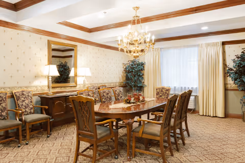 A traditional dining room with a long wooden table surrounded by upholstered chairs. The room features patterned carpet, wallpaper with a subtle design, a large mirror above a wooden sideboard with two lamps, a chandelier hanging from the ceiling, and large windows with cream-colored curtains. There are also two large potted plants in the corners.