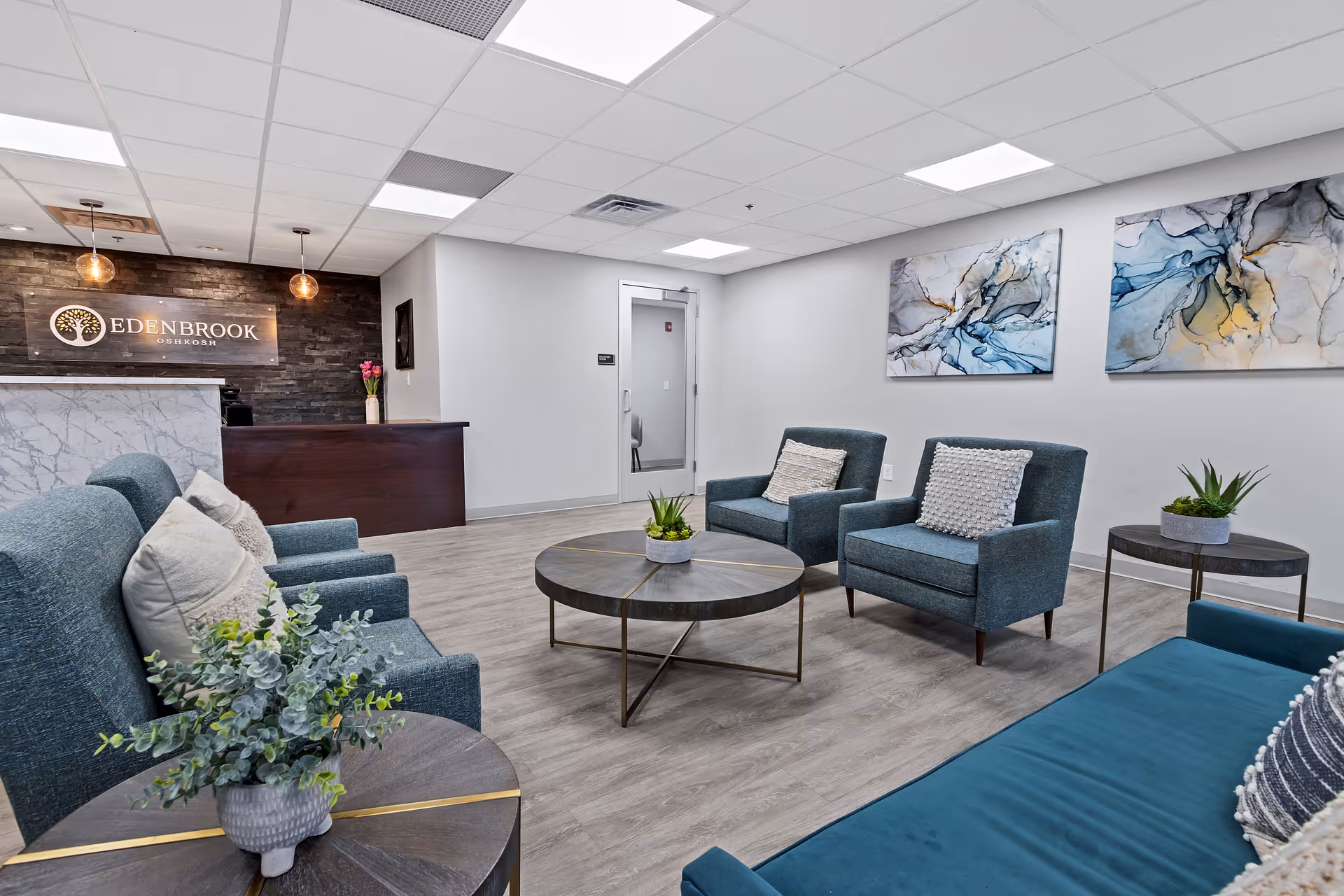 A modern and inviting reception area of Edenbrook Oshkosh featuring a dark wood reception desk with a marble front, a stone accent wall with the Edenbrook Oshkosh logo, and a seating area with blue upholstered chairs and sofas arranged around circular coffee tables with small potted plants. The room has light wood flooring, white walls, and abstract blue and gray artwork on the wall.