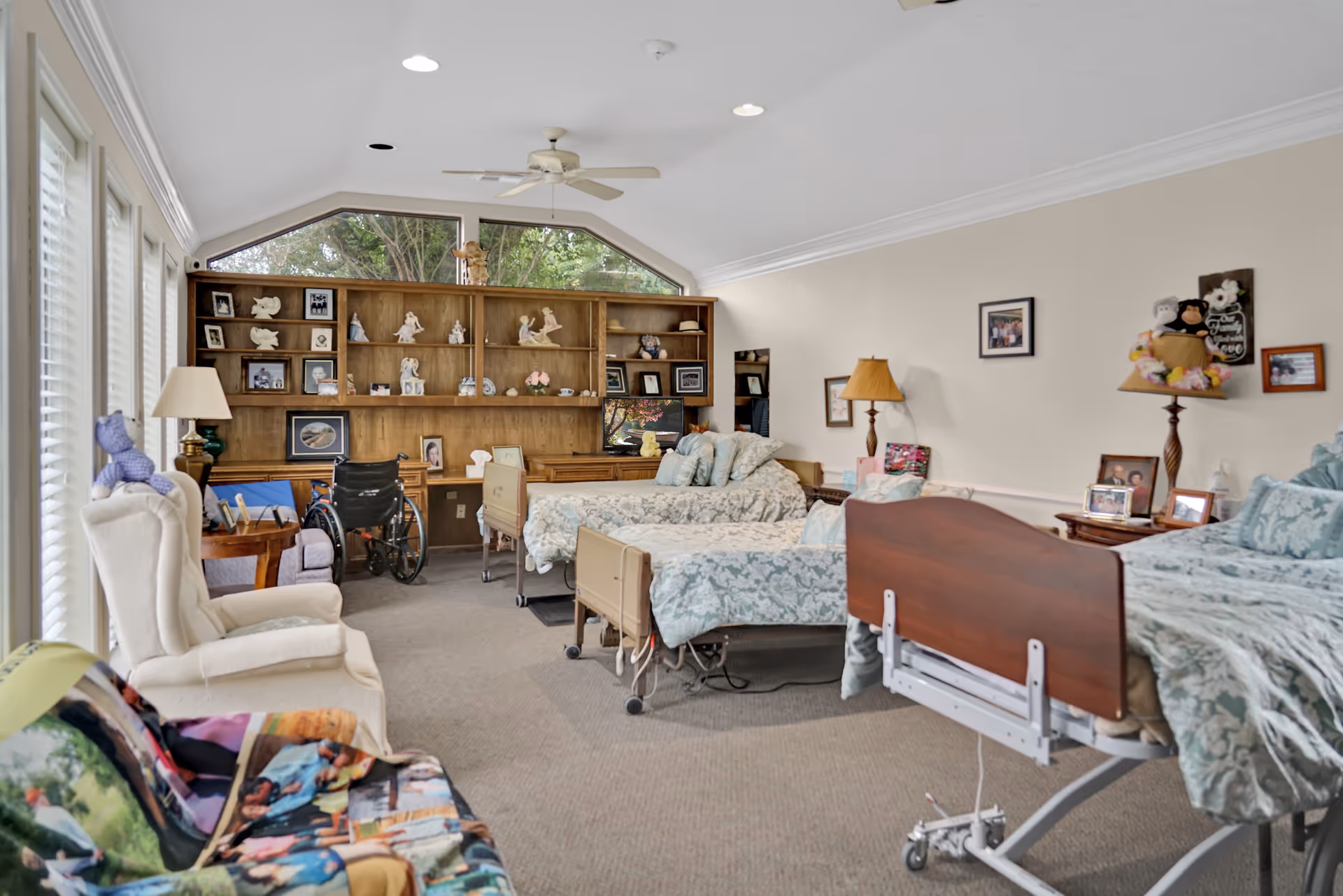 A shared bedroom in a senior living facility with three hospital-style beds, each with floral bedding. The room has a large wooden shelving unit filled with decorative items and framed photos. There are two armchairs, a wheelchair, and several lamps. The room has large windows on one side letting in natural light and a ceiling fan overhead.