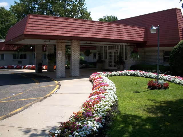 Entrance of a building with a covered driveway supported by brick pillars, surrounded by a well-maintained garden with colorful flowers lining the walkway and green grass. The building has a red roof and white brick walls.