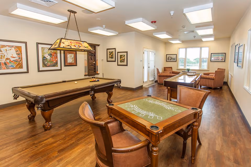 A well-lit recreational room with a billiards table, a card game table with two chairs, and a seating area with four armchairs around a coffee table. The room has wooden flooring, framed artwork on the walls, and large windows letting in natural light.