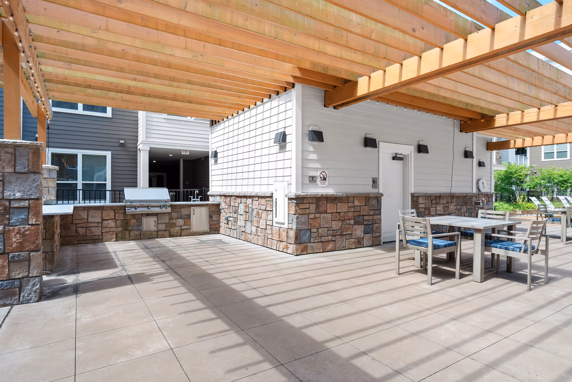 Outdoor patio area with a wooden pergola overhead casting shadows on the tiled floor. There is a built-in stone and brick grill station on the left side and a table with four chairs with blue cushions on the right. The background shows part of a building with white siding and stone accents, along with some greenery and lounge chairs further back.