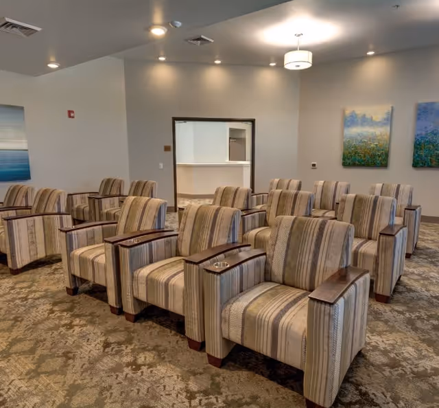 A room with multiple striped upholstered armchairs arranged in rows facing a wall with two colorful paintings. The room has beige walls, a patterned carpet, recessed ceiling lights, and a hanging light fixture. There is an open doorway leading to another room with a counter visible.