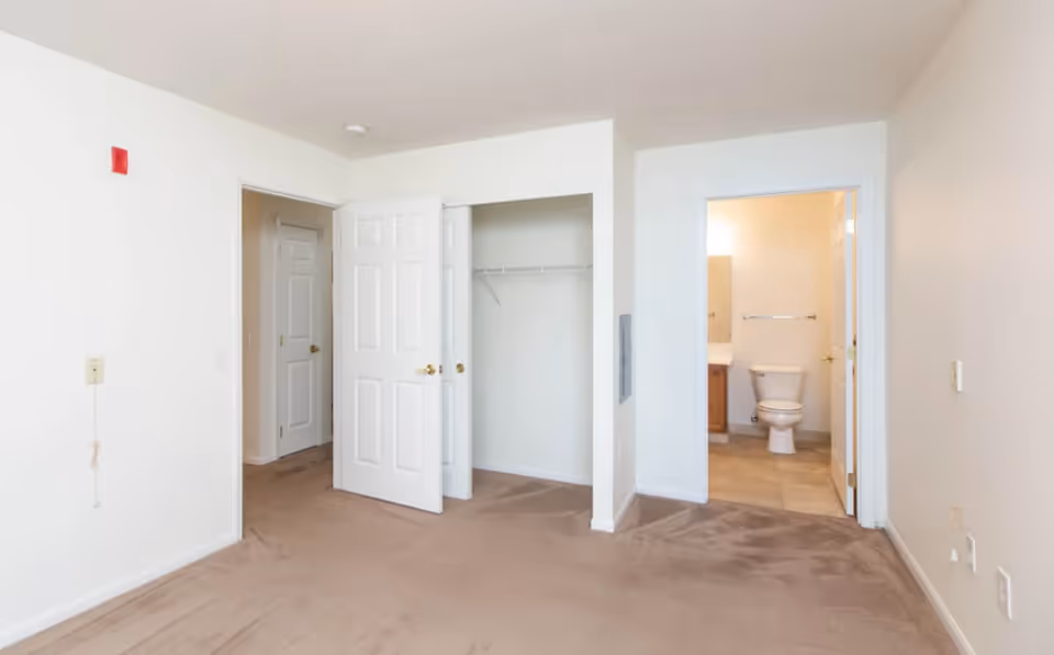 Empty carpeted bedroom with white walls, an open closet and a doorway showing an en-suite bathroom with a toilet.