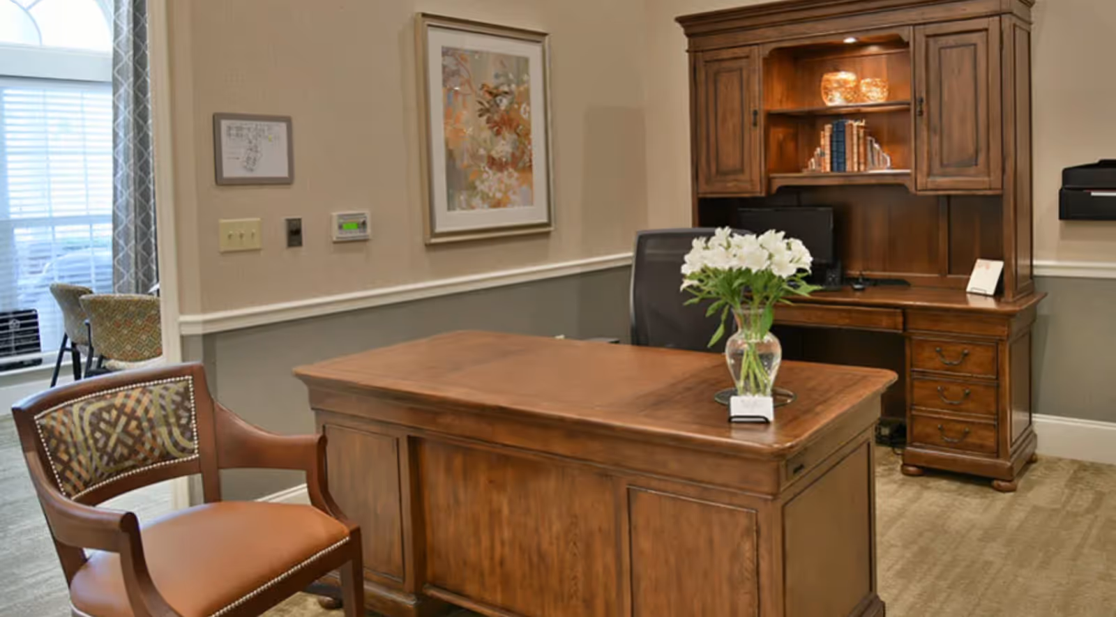 An office space with a wooden desk and matching wooden hutch. On the desk is a vase with white flowers and a small card holder. A brown leather chair with a patterned backrest is positioned in front of the desk. The wall behind the desk has a framed floral artwork and a thermostat. In the background, there is a window with blinds and a glimpse of another room with chairs and a table.