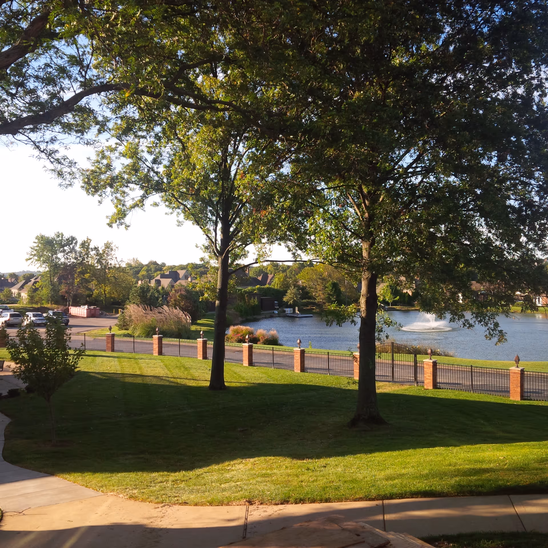 Grassy lawn with large trees overlooking a fenced pond with a fountain and houses in the background.