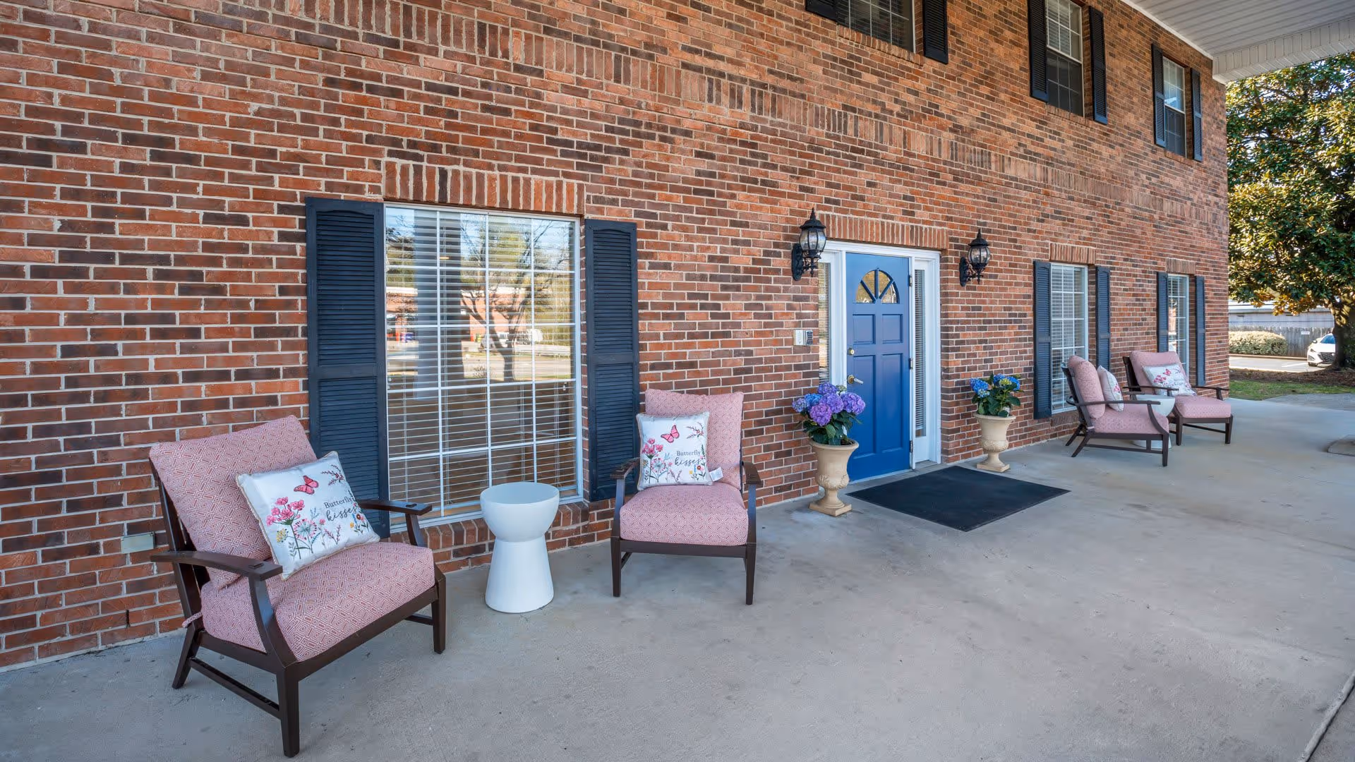 Front exterior view of a brick building with a blue door, black shutters, and large windows. There are four cushioned chairs with floral pillows arranged on the concrete porch, along with two potted plants and a small white side table.
