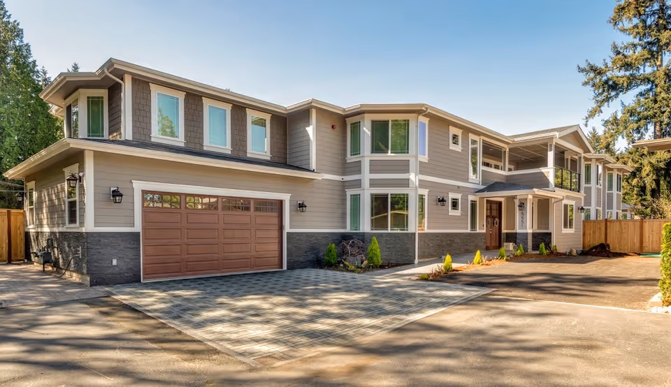 Exterior view of a two-story senior care home building with beige siding, multiple large windows, a brown garage door, and a paved driveway. The building is surrounded by trees and a wooden fence.