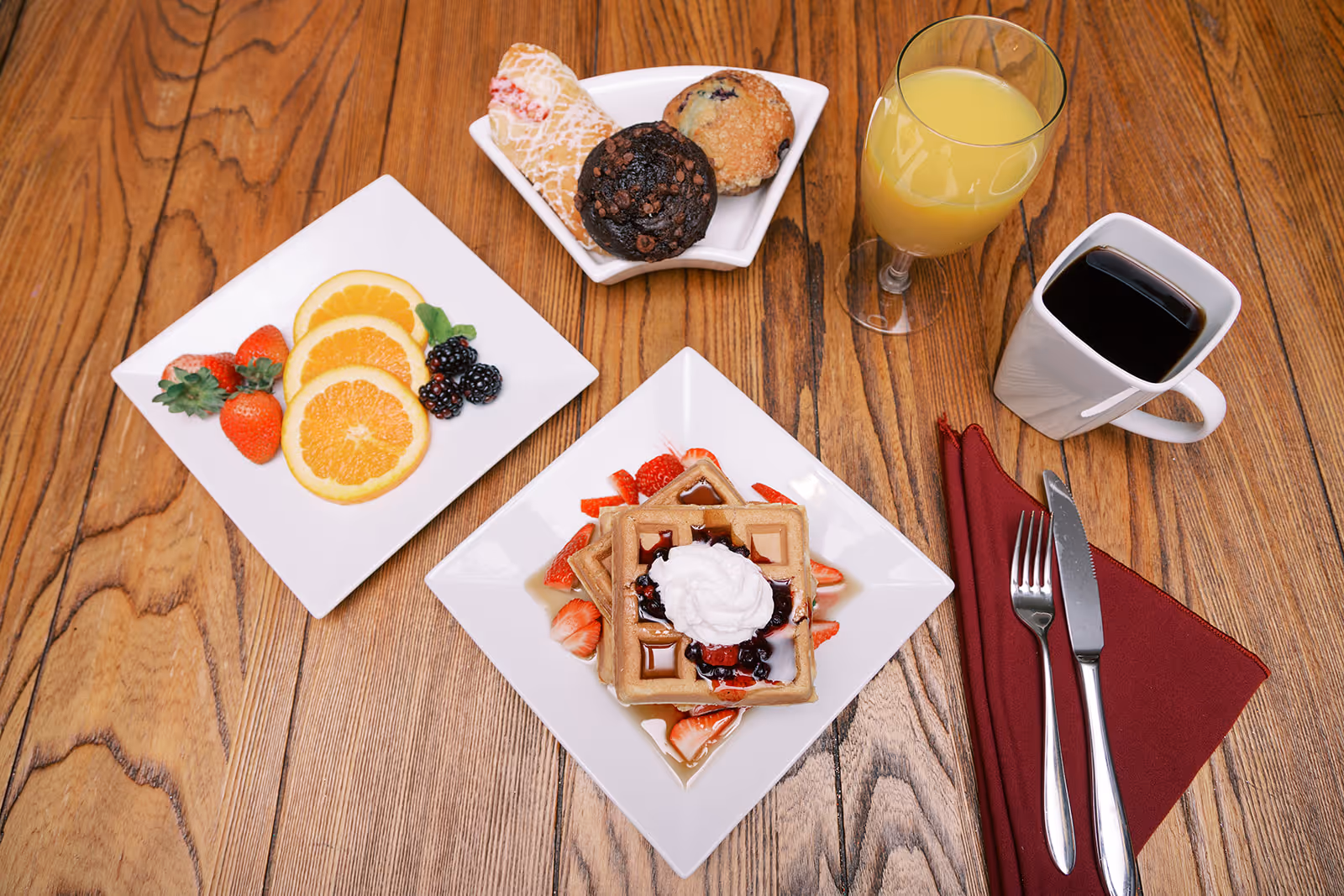 Breakfast spread on a wooden table with waffles topped with whipped cream and berries, a plate of fruit and pastries, a glass of orange juice and a cup of coffee.