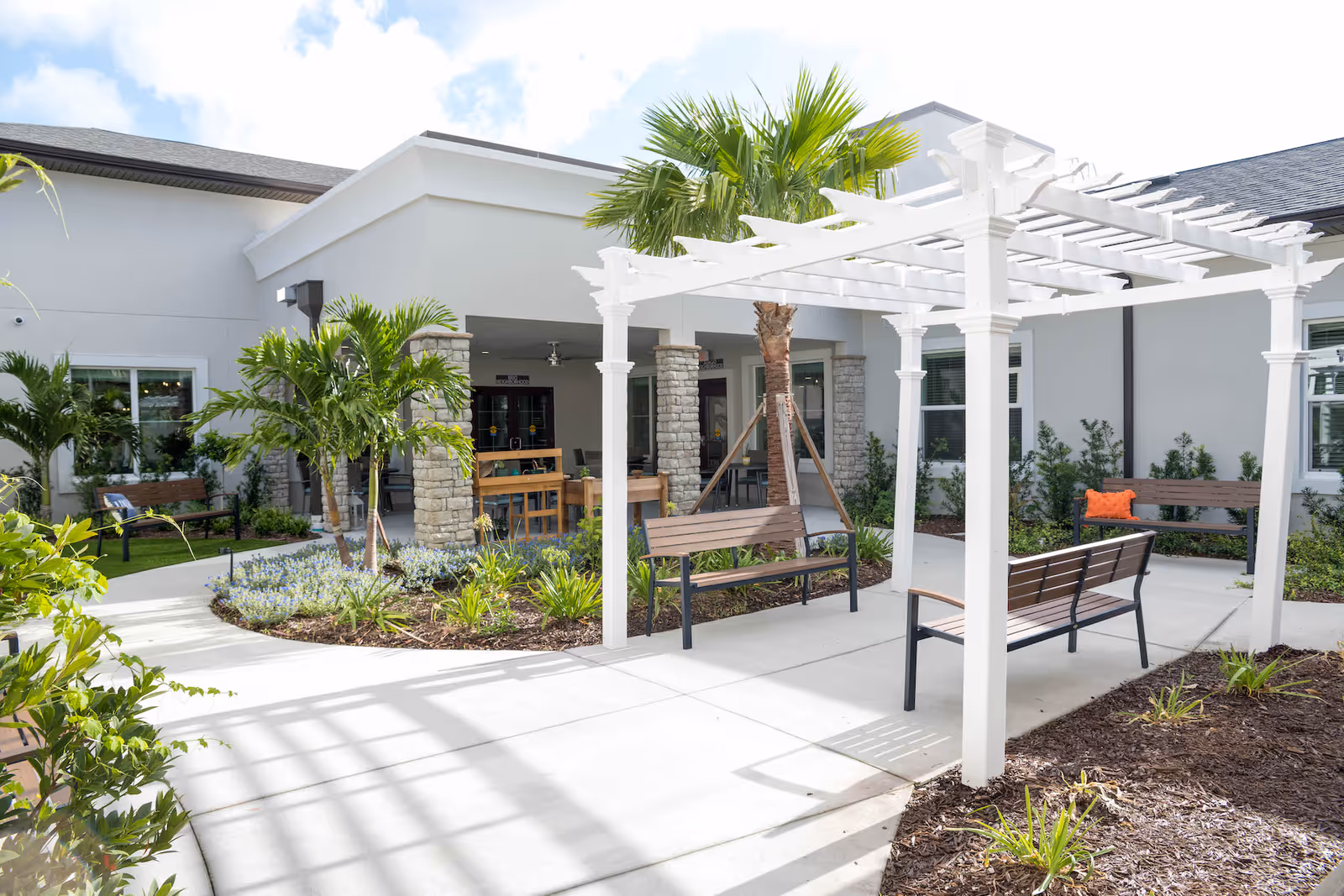 Sunny courtyard with a white pergola, benches, palm trees, and a covered seating area.