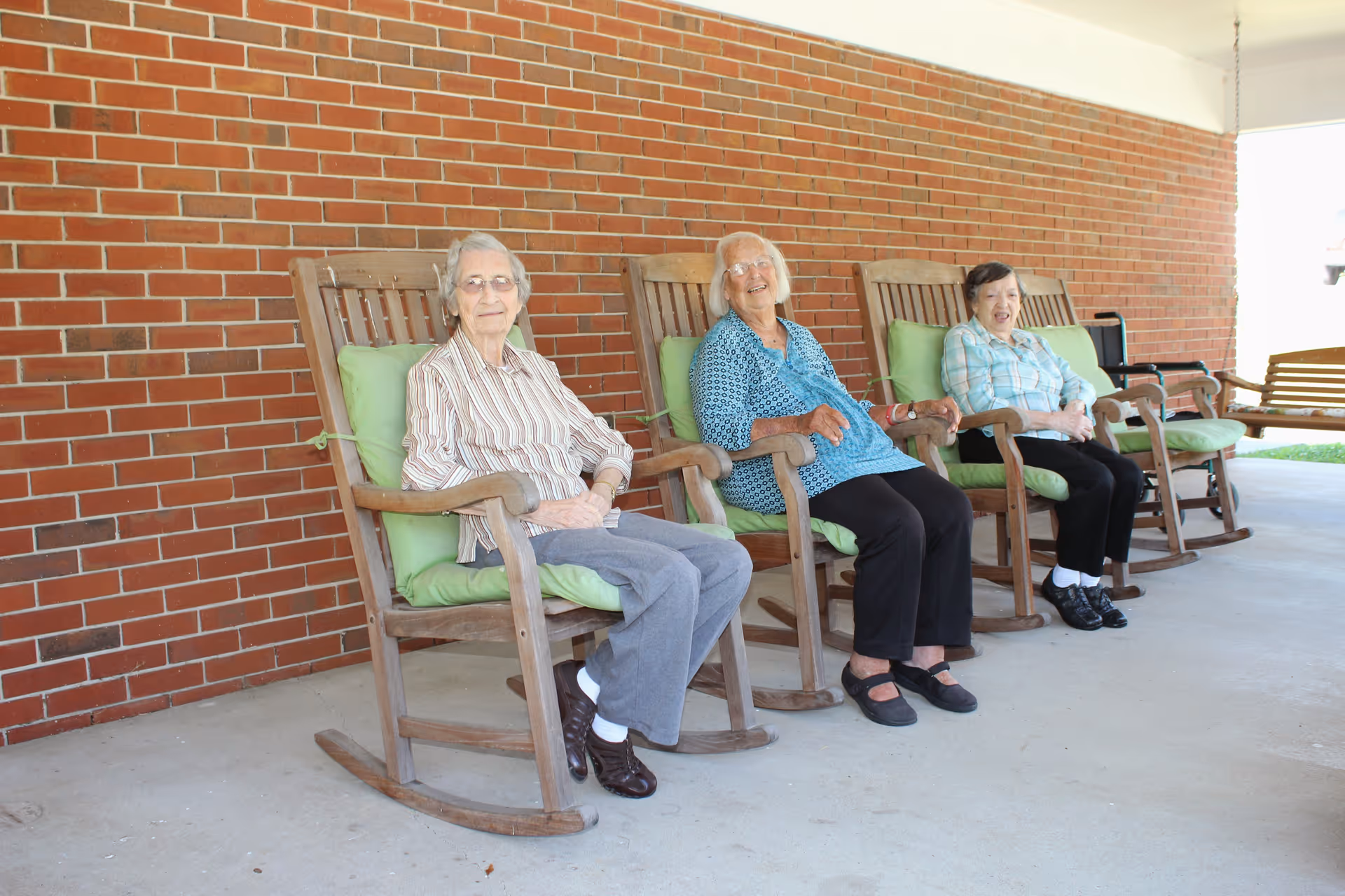 Three elderly women sitting on wooden rocking chairs with green cushions on a covered porch with a red brick wall behind them. They appear relaxed and are smiling.