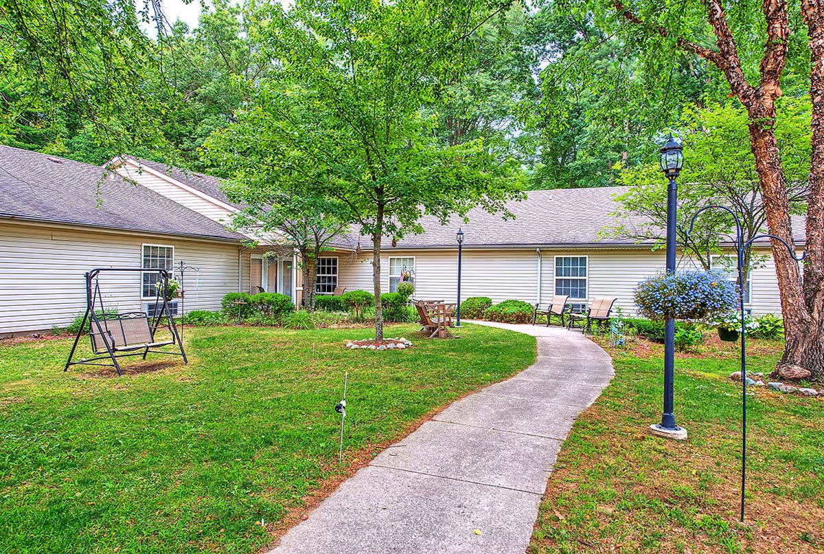 Courtyard of an assisted living facility with a curved concrete path, lawn, trees, outdoor seating, and a single-story beige building in the background.