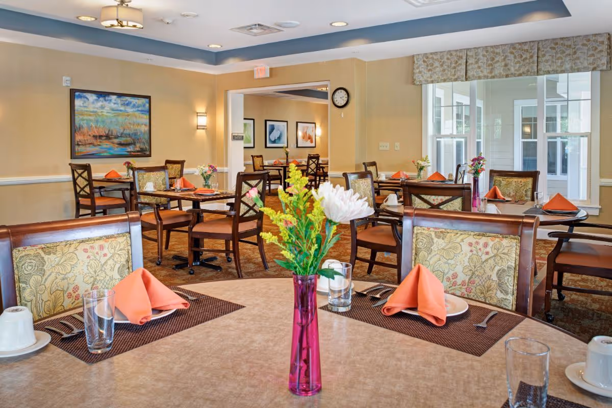 A bright and neatly arranged dining room with multiple tables set with orange napkins, plates, glasses, and silverware. Each table has a vase with colorful flowers. The room features beige walls, framed artwork, a clock, and large windows with patterned valances allowing natural light to enter.