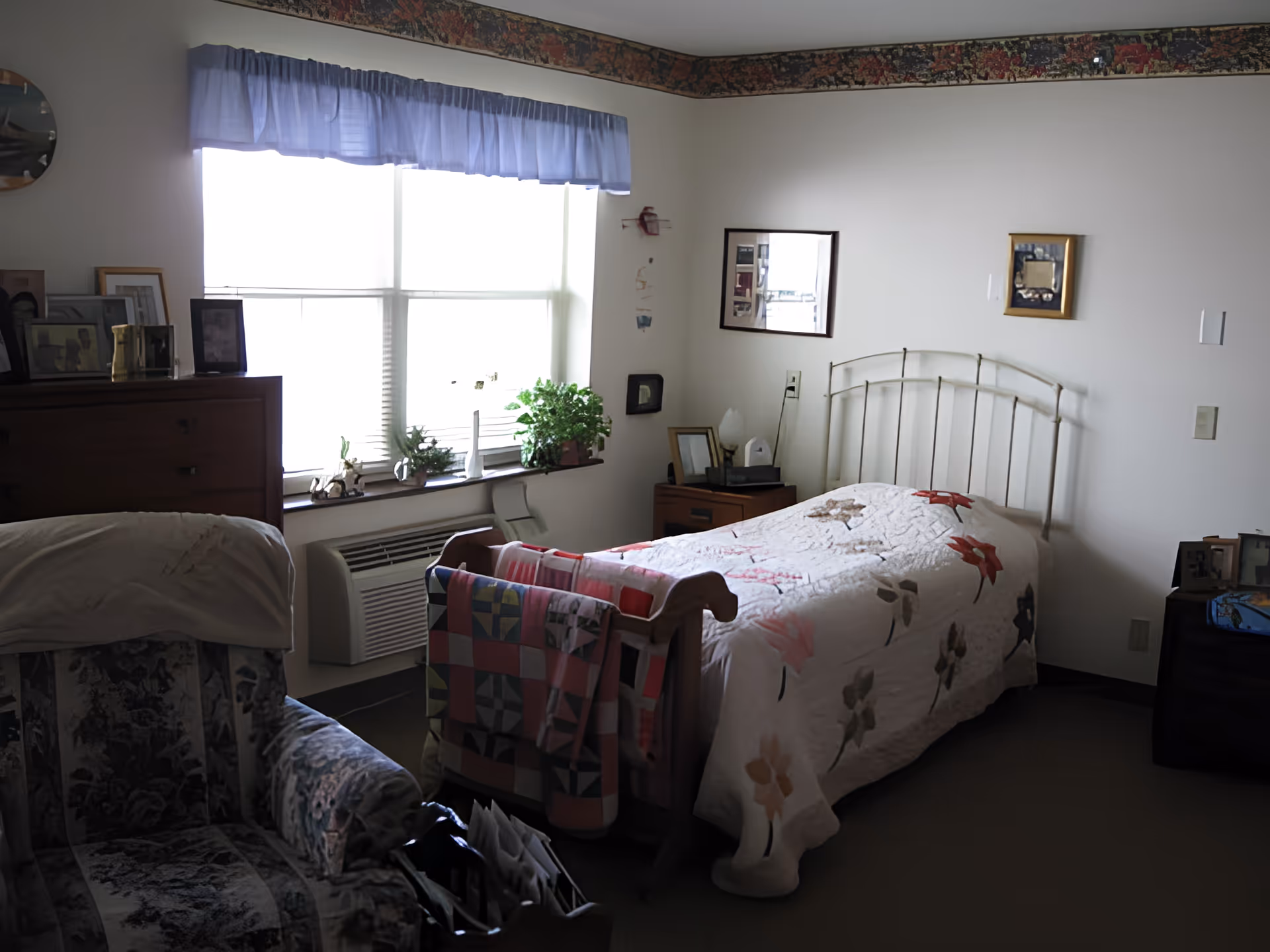 A cozy bedroom with a single bed covered in a floral quilt, a wooden quilt rack at the foot of the bed holding a colorful patchwork quilt, a floral upholstered armchair, a wooden dresser with framed photos, and a window with blue curtains letting in natural light. There are small plants on the windowsill and framed pictures on the walls.