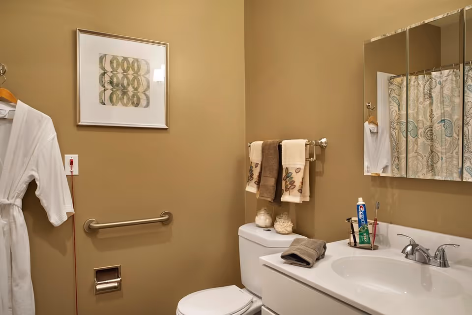 A bathroom with beige walls featuring a white toilet, a white sink with a silver faucet, and a mirrored medicine cabinet. There are three towels hanging on a towel rack above the toilet, a white bathrobe hanging on the wall, a framed abstract artwork, and a shower curtain with a paisley pattern. Various toiletries including toothpaste and a toothbrush are on the sink counter.