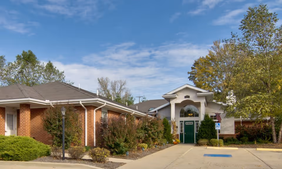 Exterior view of a single-story brick building with a green door entrance, surrounded by trees and shrubs under a partly cloudy blue sky. There is a sidewalk leading to the entrance and a handicap parking space visible in front.