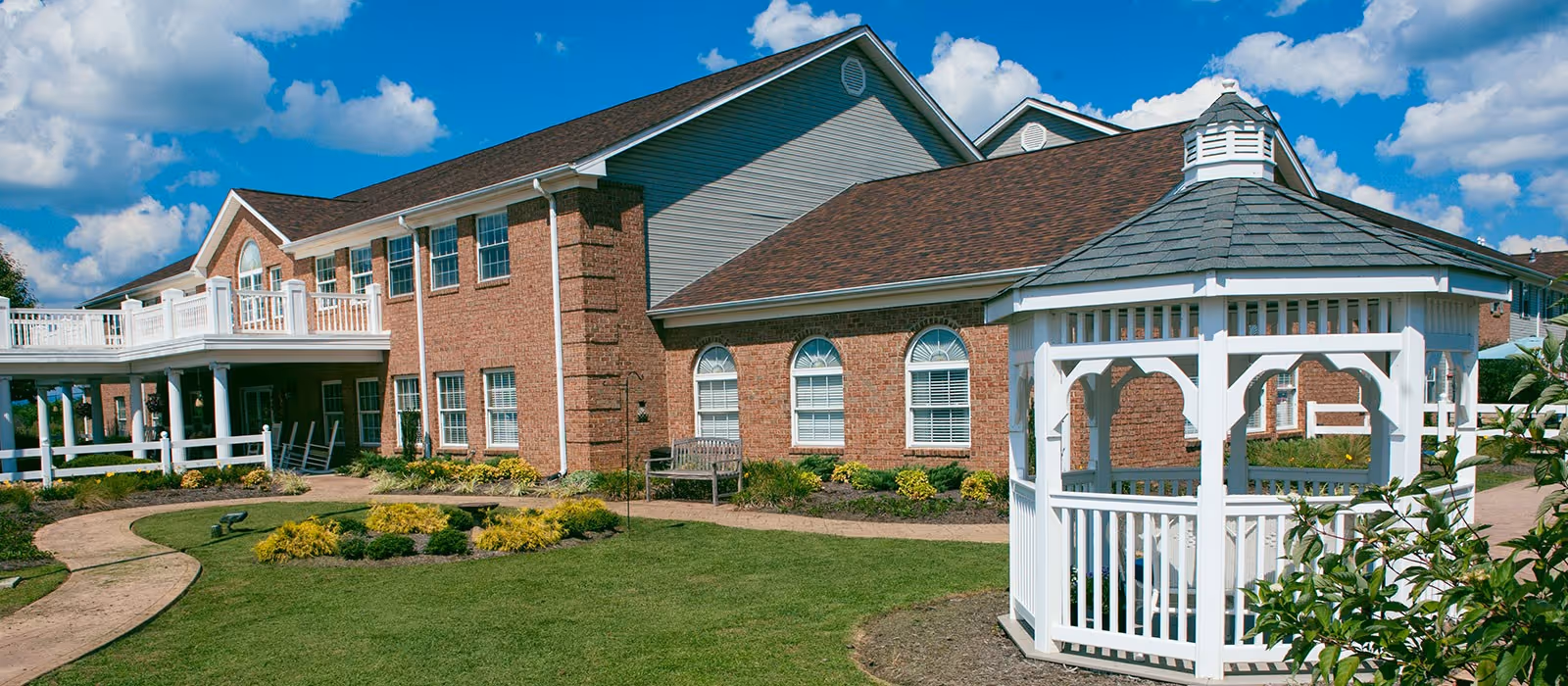 Exterior view of Commonwealth Senior Living at Christiansburg showing a two-story brick building with multiple windows, a white balcony, a well-maintained garden with shrubs and a curved walkway, and a white gazebo in the foreground under a partly cloudy blue sky.
