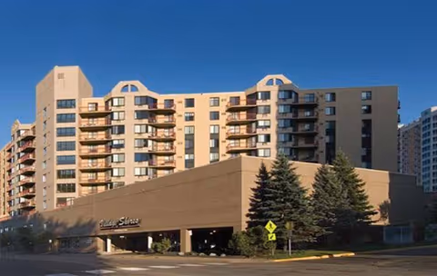 Exterior view of a multi-story senior living facility named Village Shores with balconies, large windows, and a parking garage at the base. There are trees and a pedestrian crossing sign in front of the building under a clear blue sky.
