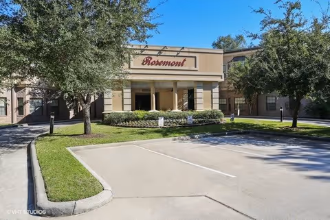 Front exterior view of the Rosemont Assisted Living And Memory Care building with a driveway and parking spaces in front, surrounded by trees and landscaping under a clear blue sky.
