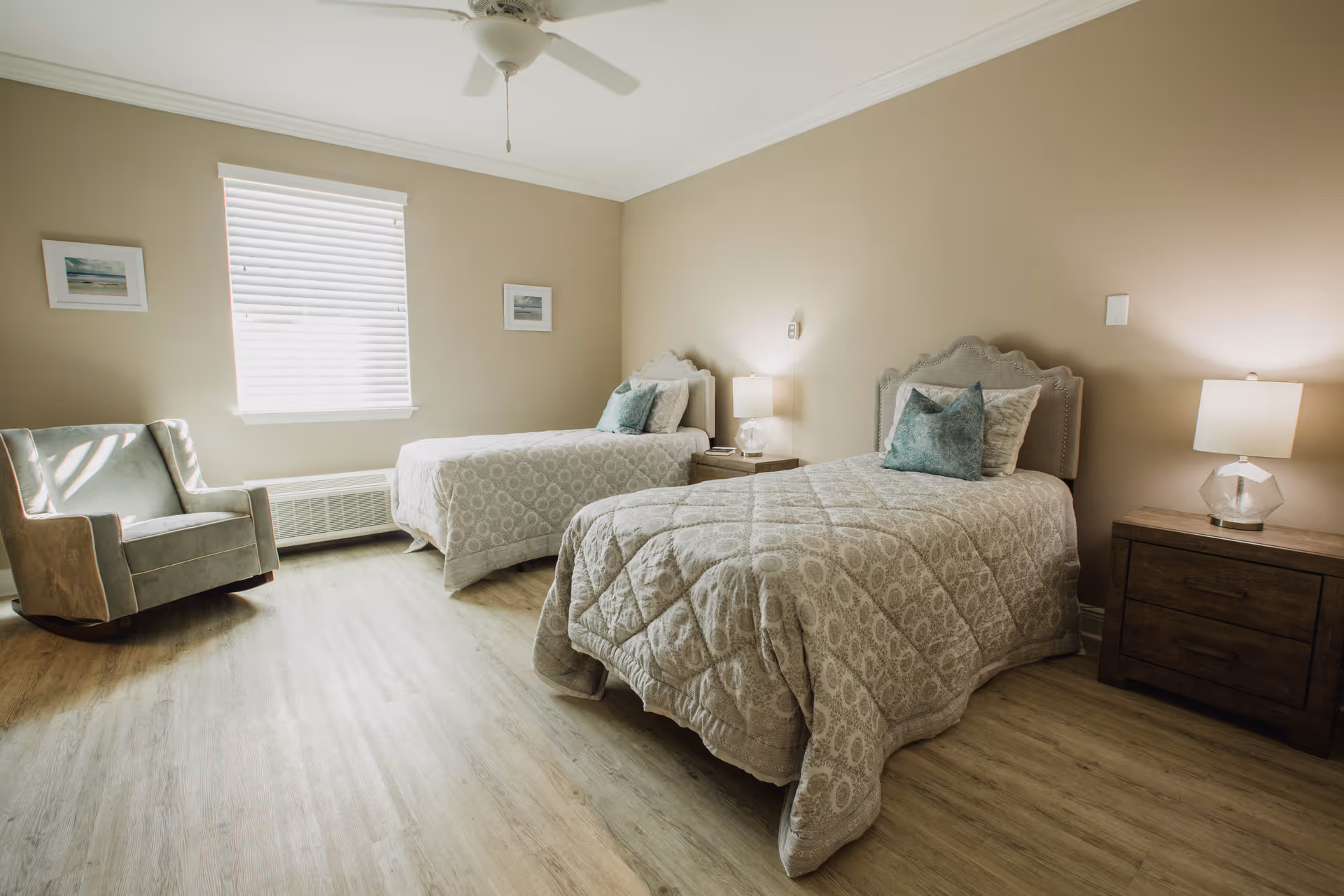 A bright bedroom with two twin beds, each with a patterned beige comforter and a teal pillow. Between the beds is a wooden nightstand with a lamp. There is a window with blinds on the left wall, a light blue armchair beneath it, and two framed pictures on the walls. The floor is light wood, and a ceiling fan is visible above.