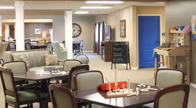 A spacious common area in a senior living facility with multiple tables and chairs arranged for activities. One table has a ring toss game set up. The room has beige walls, a bright blue door, and a chalkboard sign in the background. There are various pieces of furniture including couches and a globe, with soft lighting from ceiling fixtures.