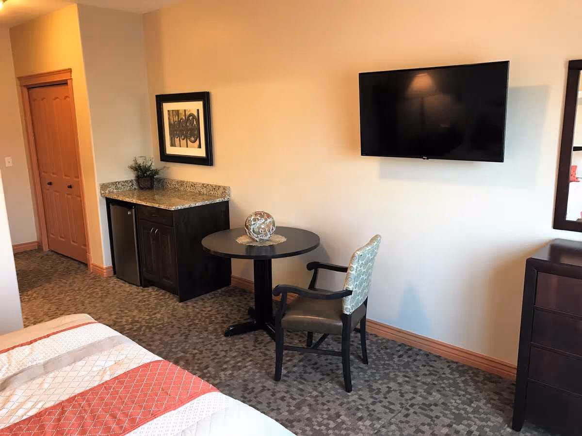 Small assisted-living bedroom with a bed, wall-mounted TV, round table and chair, and a countertop with a mini-fridge.