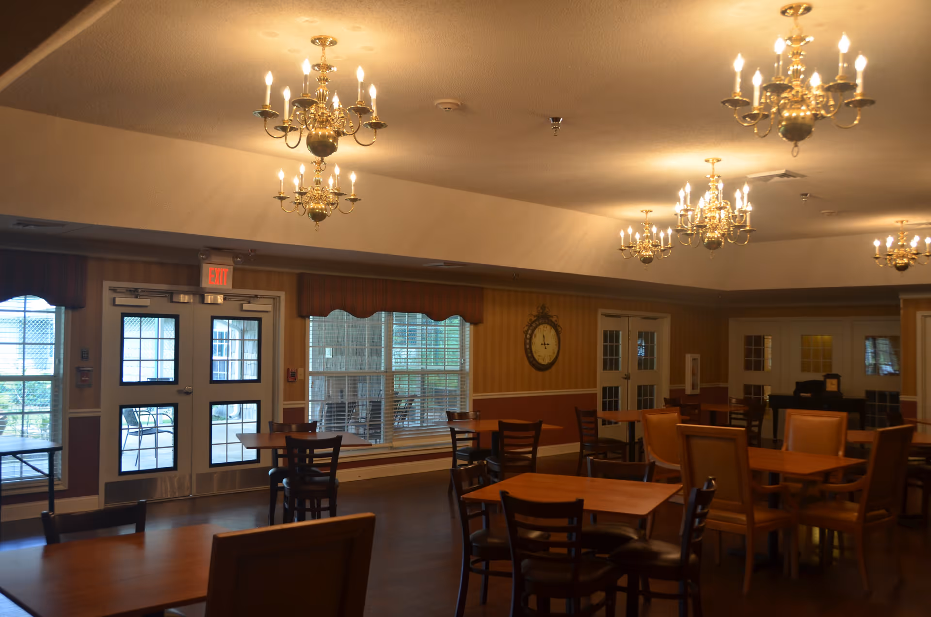 Interior view of a dining room with multiple wooden tables and chairs arranged neatly. The room features several ornate chandeliers hanging from the ceiling, a large clock on the wall, and windows with valances. Double doors and an exit door lead to an outdoor area with patio furniture visible through the windows.