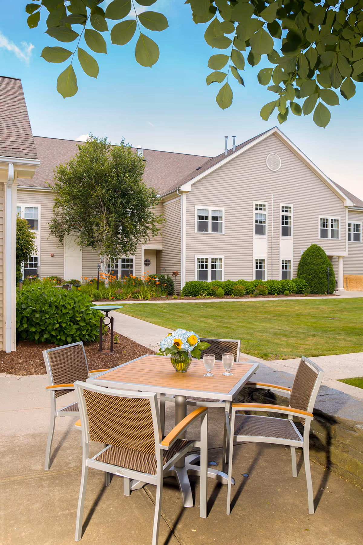 Patio table with four chairs and a vase of flowers on a courtyard in front of a beige multi-unit senior living building.