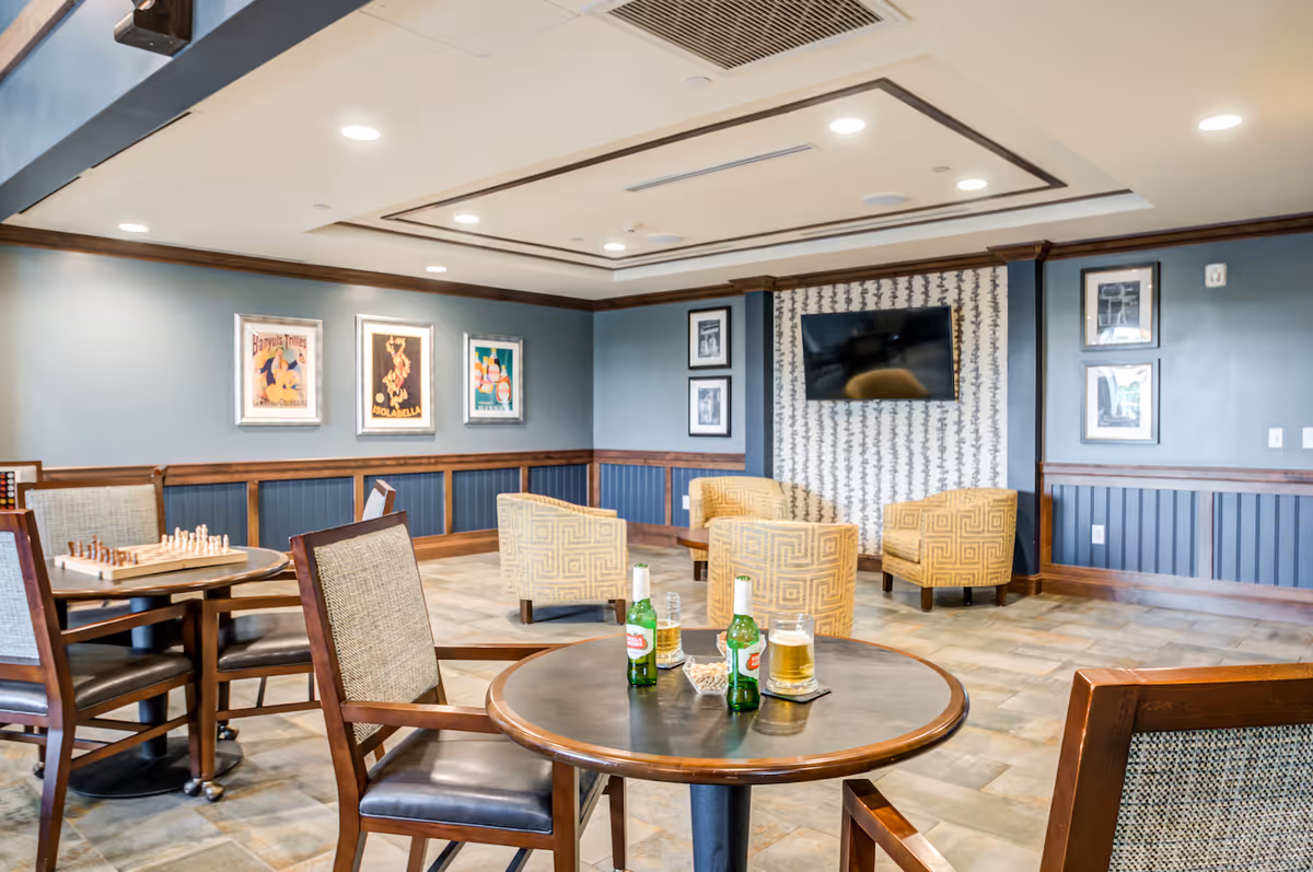 A cozy common area in a senior living facility featuring a round table with chairs, two bottles of beer and two glasses of beer on the table. In the background, there are four upholstered armchairs arranged around a wall-mounted flat screen TV. The walls are decorated with framed artwork and photographs, and the room has a warm, inviting atmosphere with recessed lighting and wood trim.