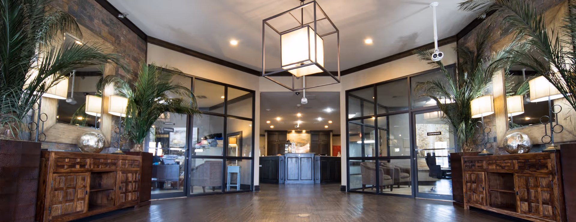 Spacious and well-lit interior common area with wooden flooring, large mirrors on both sides, tall green plants in pots, wooden cabinets, and glass-walled rooms visible in the background. A modern square pendant light hangs from the ceiling.