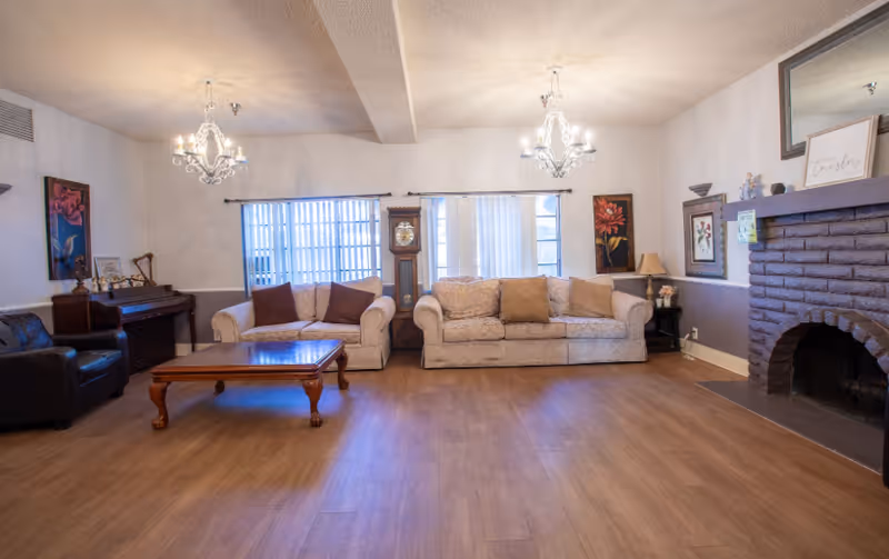 A cozy living room with two beige sofas adorned with brown cushions, a wooden coffee table in the center, a dark leather armchair, and a piano against the left wall. The room features two chandeliers hanging from the ceiling, a grandfather clock between two windows with vertical blinds, and a brick fireplace on the right side decorated with framed pictures and small ornaments.