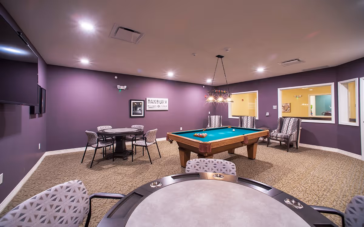 Recreation room with a pool table at center, round game tables, and upholstered chairs against purple walls.