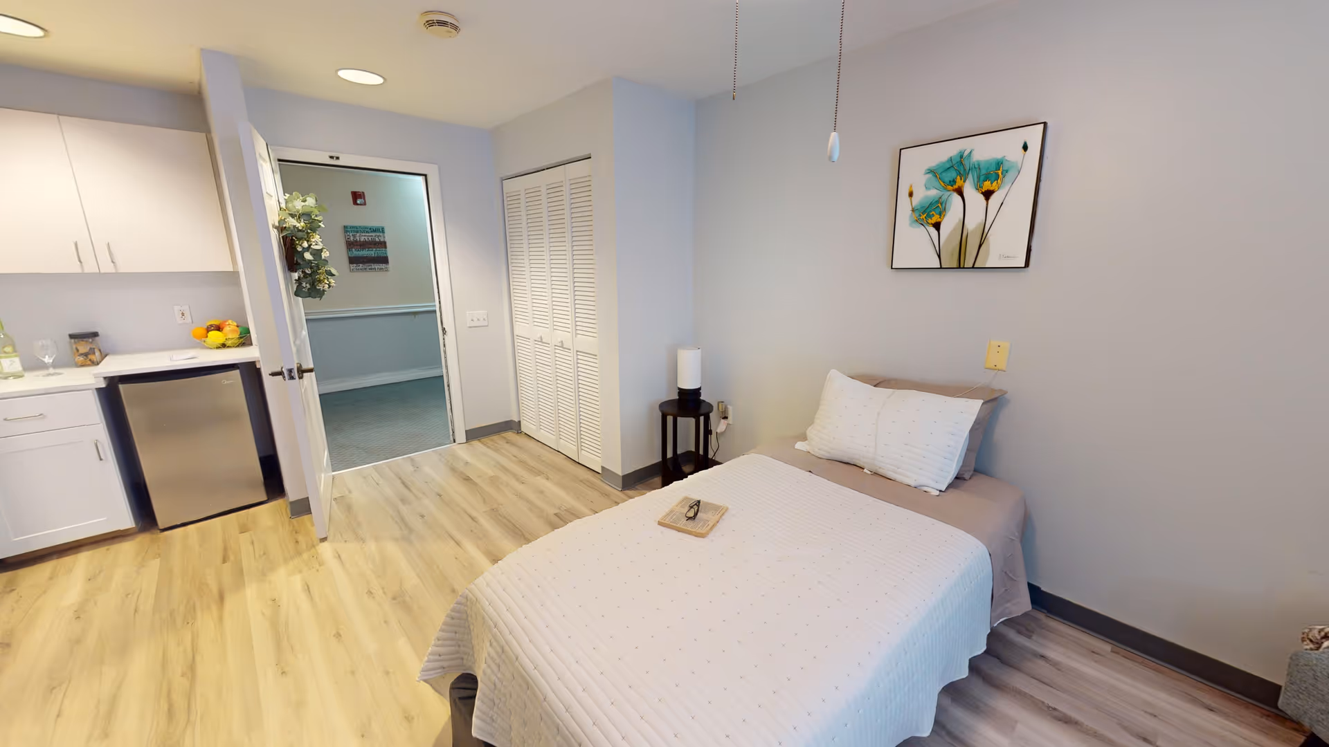 A small, clean bedroom with a single bed covered in a white quilt and two pillows. A book and glasses rest on the bed. To the left, there is a kitchenette area with white cabinets, a small refrigerator, and a countertop with fruit and a bottle. The room has light wood flooring, a closet with white louvered doors, and a door leading to a hallway. A colorful floral painting hangs on the wall above the bed.