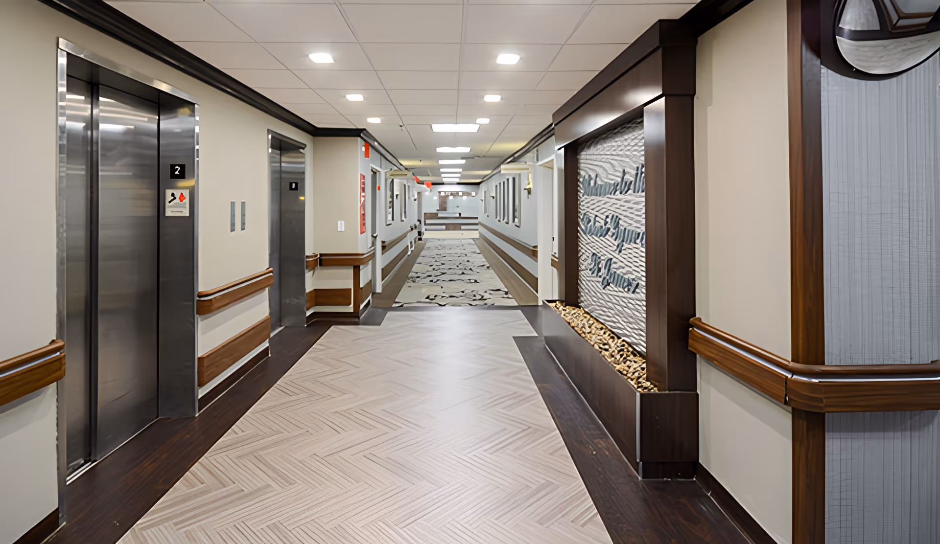 A clean, well-lit hallway in a healthcare facility with two stainless steel elevators on the left side and a decorative wall feature on the right that reads 'St. James Rehabilitation & Healthcare Center'. The hallway has light-colored flooring with a patterned carpet further down and wooden handrails along the walls.