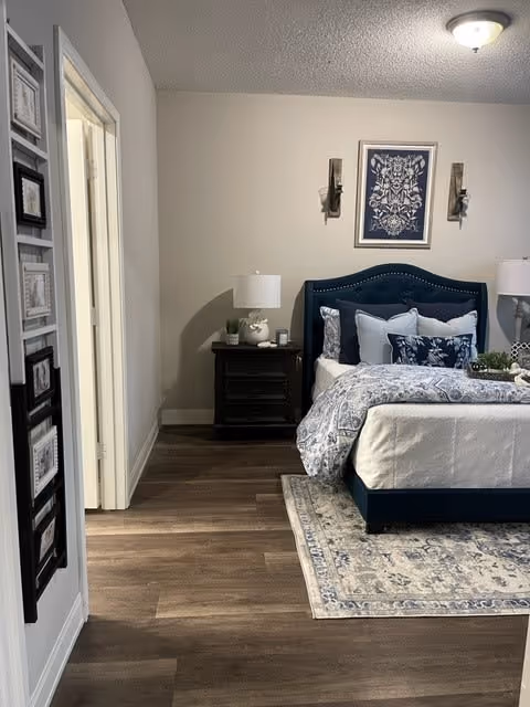 A neatly made bed with a blue upholstered headboard and multiple pillows in shades of blue and white. The bed is placed on a patterned area rug with blue and beige tones. On either side of the bed are dark wooden nightstands with white lamps. Above the bed hangs a framed piece of artwork with a floral design. The room has wood flooring and light-colored walls. To the left, there is an open doorway leading to another room, and a wall-mounted photo frame is visible on the left wall.
