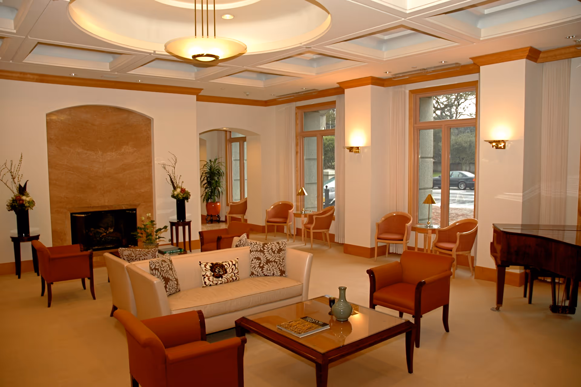 A spacious and elegant living room with a beige sofa adorned with patterned cushions, surrounded by four brown armchairs. A wooden coffee table with decorative items sits in the center. The room features large windows with natural light, a grand piano on the right, a fireplace with a marble surround, and floral arrangements on small tables. The ceiling has a coffered design with a central light fixture.