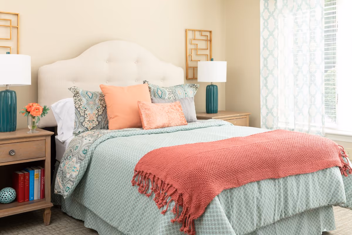 A neatly made bed with a cream-colored tufted headboard, patterned pillows in shades of green and coral, and a coral knitted throw blanket at the foot. There are two wooden nightstands on either side of the bed, each with a teal lamp and one with a small vase of orange flowers. Light filters in through a window with sheer patterned curtains on the right side of the room.