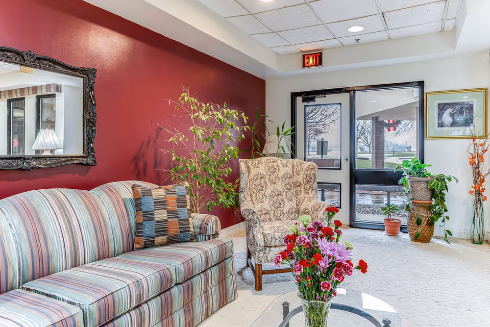 A cozy seating area in a senior living facility with a striped sofa, a patterned armchair, a glass coffee table with a vase of colorful flowers, several potted plants, a large ornate mirror on a red accent wall, and a glass door with an exit sign above it.