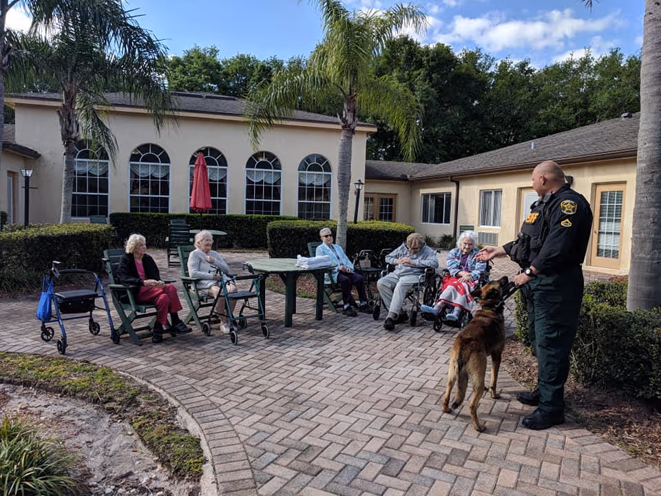 A group of elderly people sitting outside on chairs and wheelchairs in a courtyard area with palm trees and a building in the background. A uniformed officer is standing in front of them with a police dog. The sky is partly cloudy.