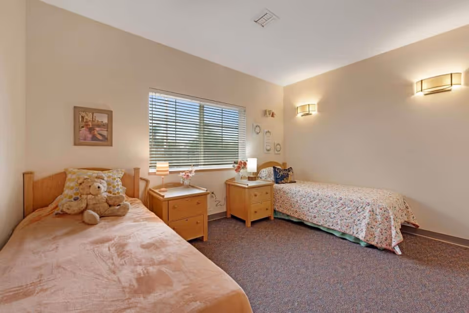 Two-bed furnished bedroom with nightstands, lamps, a window with blinds, and a teddy bear on one bed.