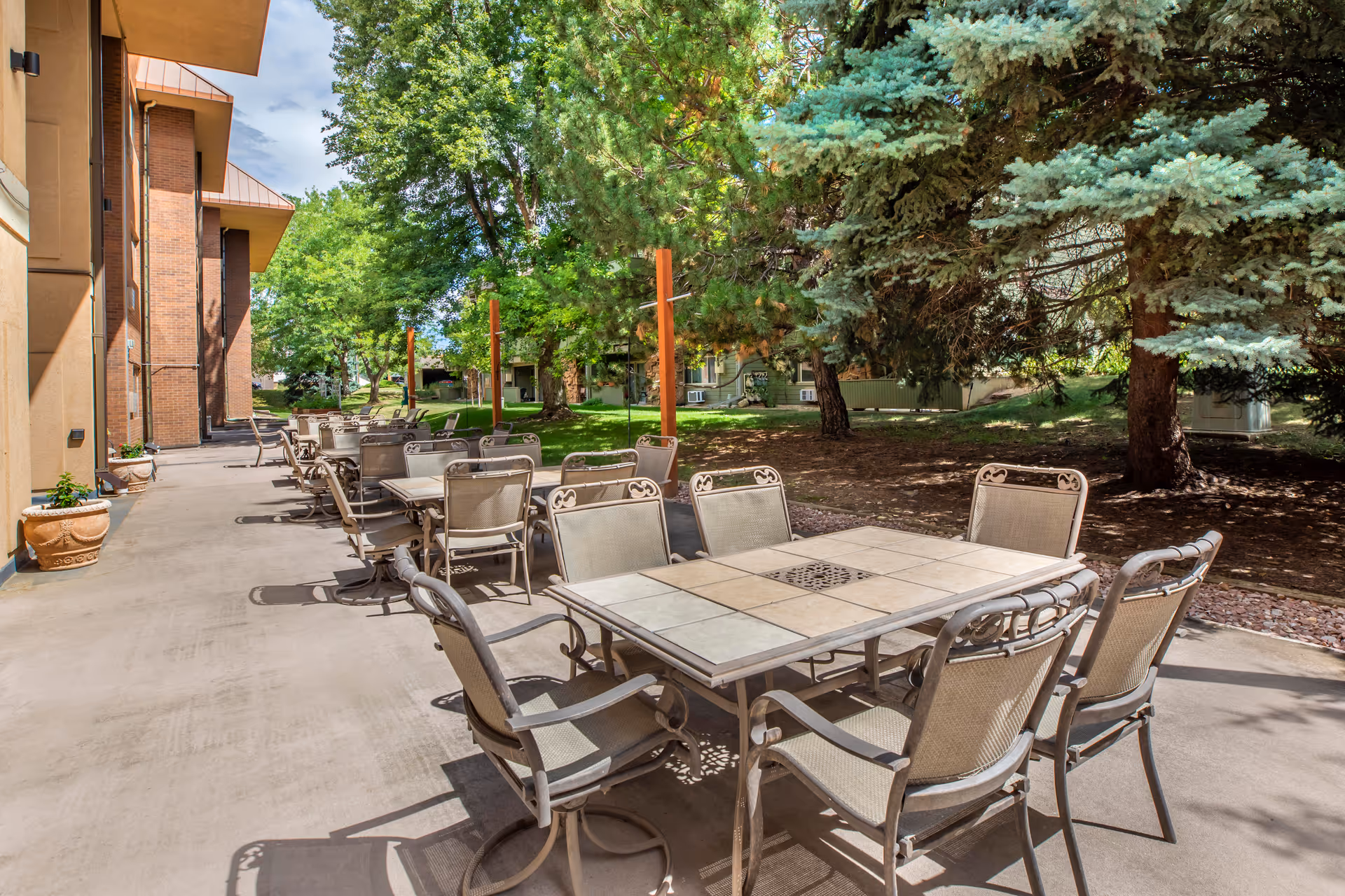 Outdoor patio area at Brookdale North Boulder with multiple metal tables and chairs arranged on a concrete surface. The patio is adjacent to a building and surrounded by green trees and grass under a partly cloudy sky.