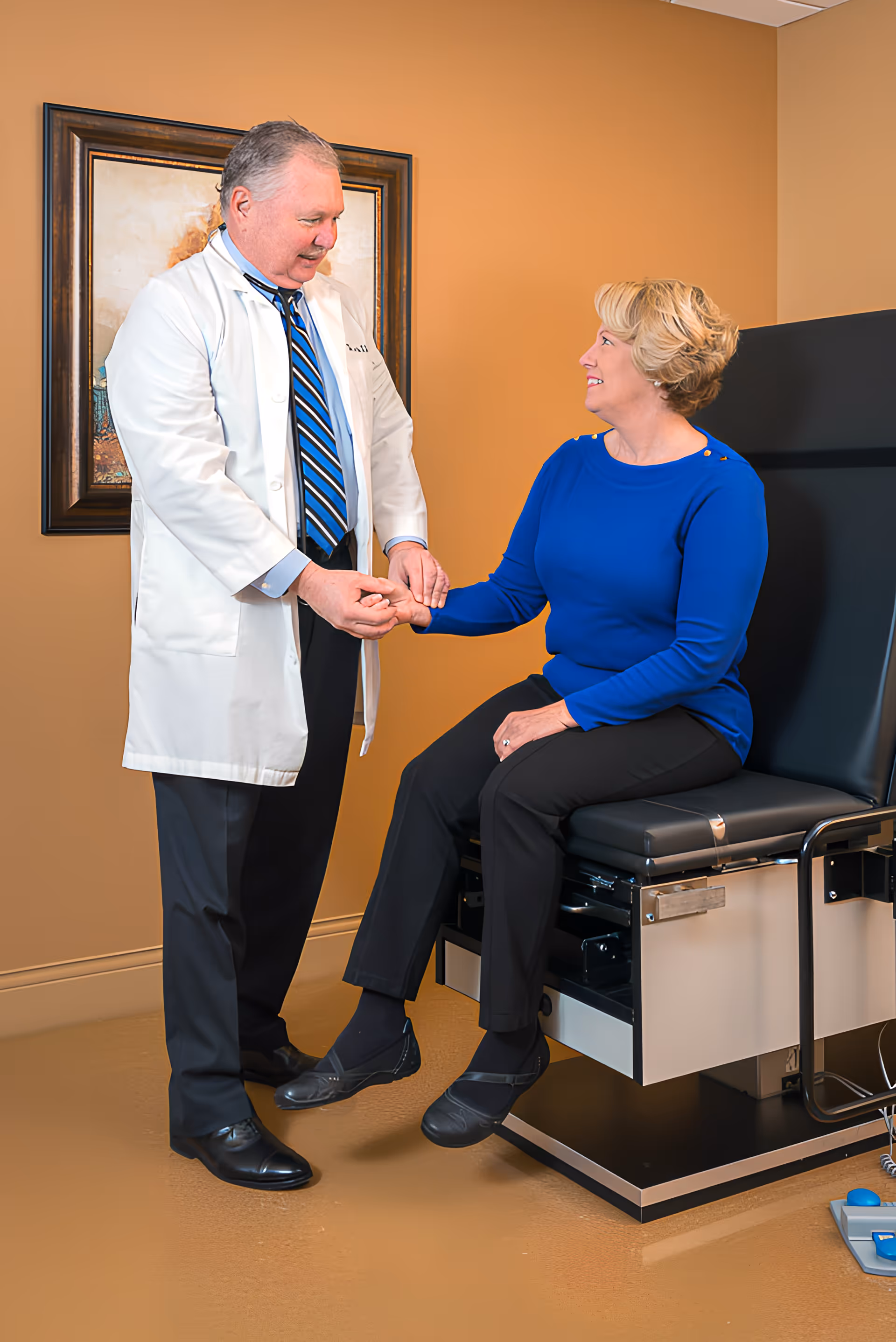 A doctor in a white coat is holding the hand of a seated woman wearing a blue top and black pants in a medical examination room with beige walls and a framed picture on the wall.
