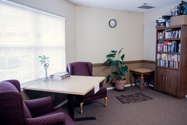 A cozy reading nook featuring two purple armchairs, a small table, a potted plant, and a bookshelf filled with books, illuminated by natural light from a window.