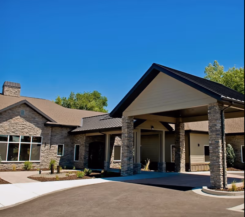 Exterior view of a single-story assisted living facility building with stone and beige siding, large windows, and a covered entrance supported by stone pillars under a clear blue sky.