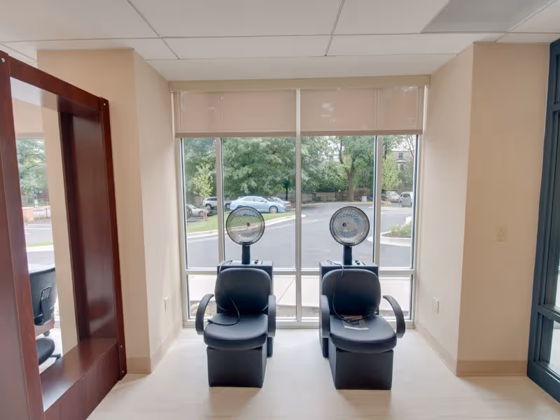 Two black salon hair dryer chairs positioned in front of large windows with a view of parked cars and trees outside. A large mirror is mounted on the left wall, and the room has beige walls and a light-colored floor.