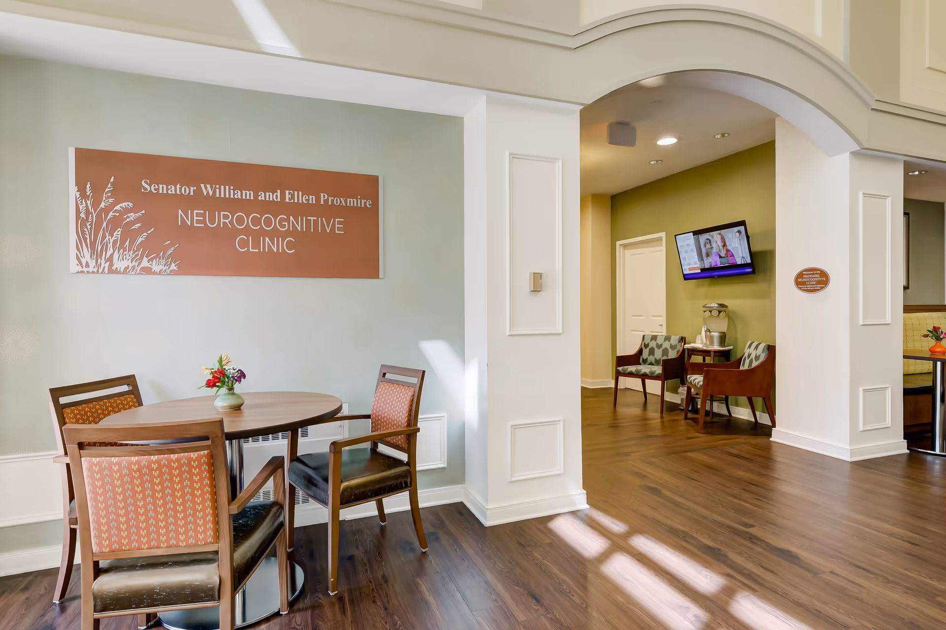 Interior view of the Senator William and Ellen Proxmire Neurocognitive Clinic featuring a round wooden table with four chairs, a small flower vase on the table, wood flooring, a green accent wall with two chairs and a water dispenser, and a wall-mounted TV.