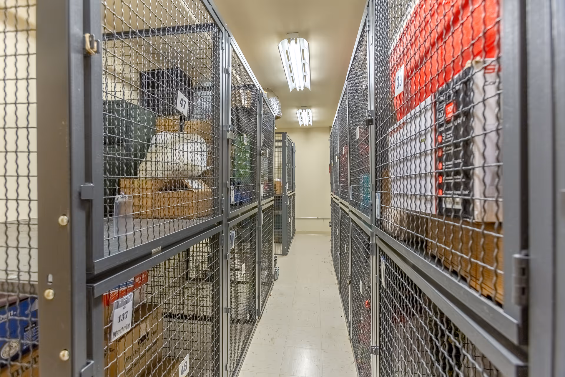 Narrow corridor lined with numbered metal storage cages holding boxes and personal belongings on both sides.