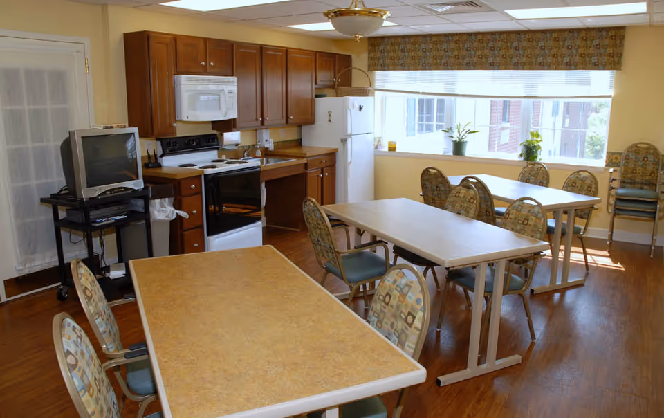 A bright room with wooden flooring featuring a small kitchen area with wooden cabinets, a white refrigerator, stove, microwave, and sink. In front of the kitchen are three tables with patterned chairs around them. A large window with a valance lets in natural light, and a TV on a stand is positioned near a door with a sheer curtain.